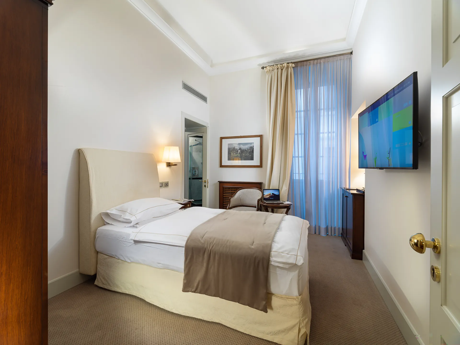 Nell'immagine, la camera singola del Grand Hotel della Posta: si vede il letto singolo e, di fronte, la TV appesa alla parete. In fondo alla stanza, di fianco alla finestra, un tavolino tondo con una poltrona. Sul tavolino è poggiato un laptop acceso.