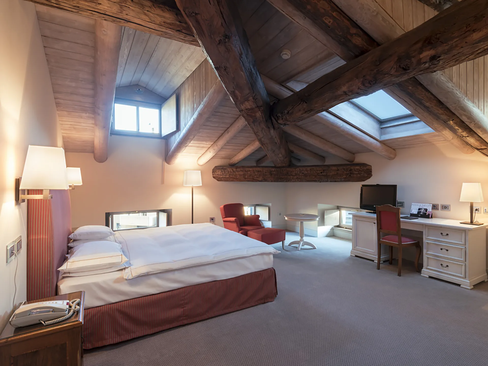Nell'immagine, la camera Superior mansardata del Grand Hotel della Posta: si vede il letto matrimoniale sulla sinistra e, di fronte, un tavolo con la TV. Sulla sinistra, una poltroncina con un tavolino tondo. Sul soffitto, il lucernario e le travi in legno a vista.